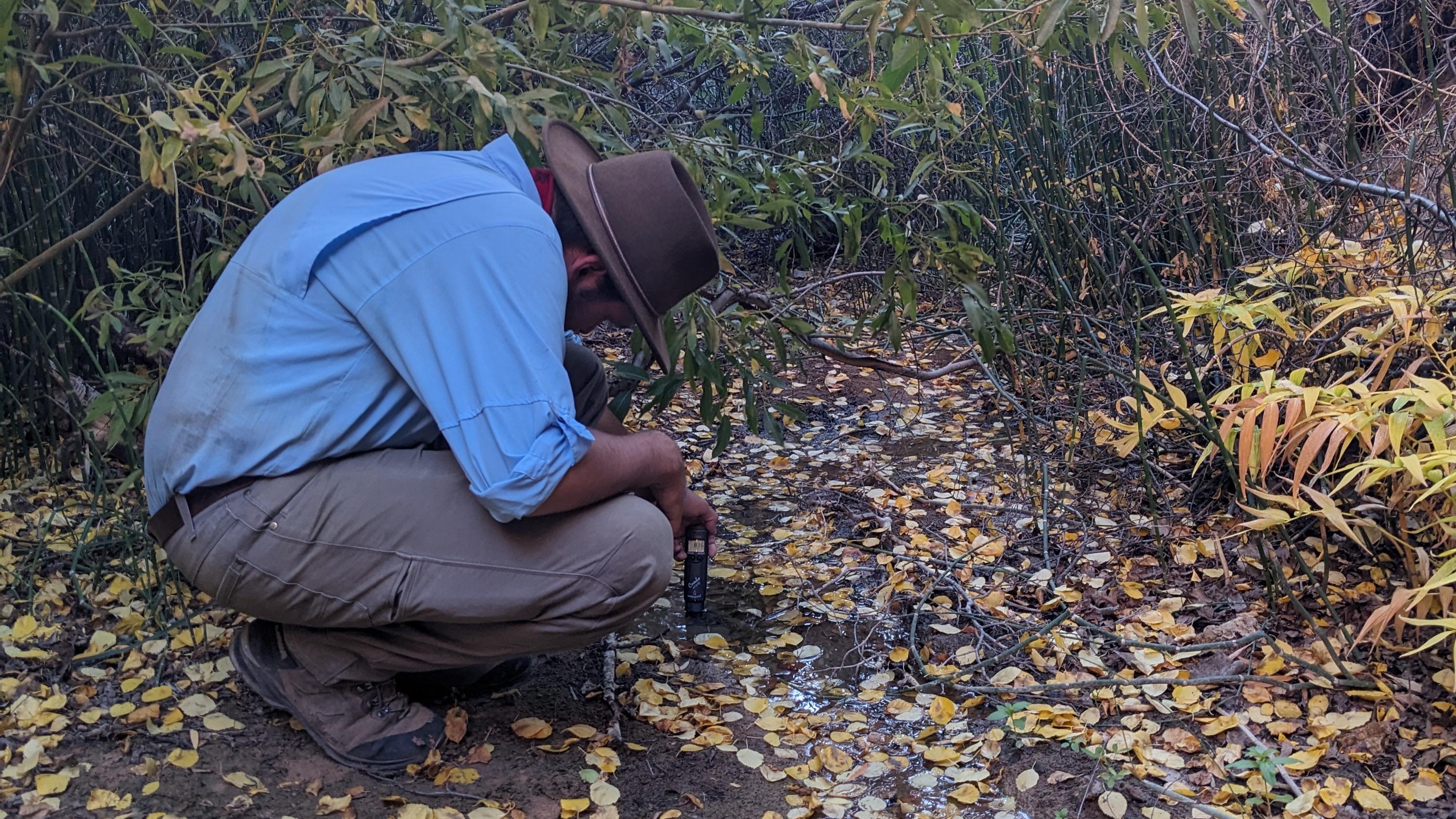 person in blue shirt and brown pants kneels over a creek