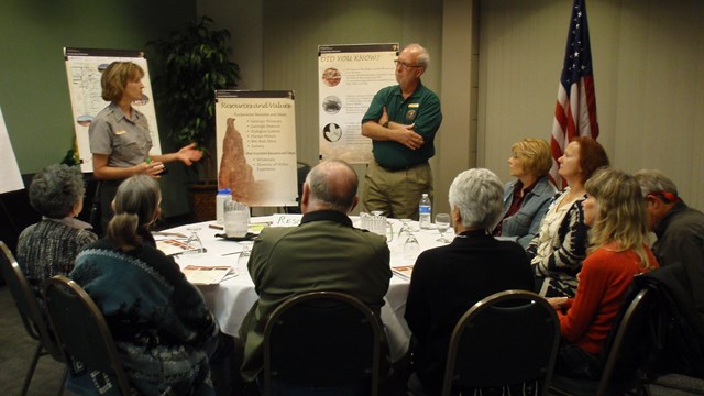 park ranger and volunteer present to a round table of others in a dimly lit room