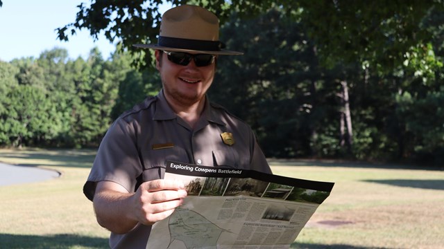 A park ranger stands in the shade holding a brochure and wearing sunglasses 