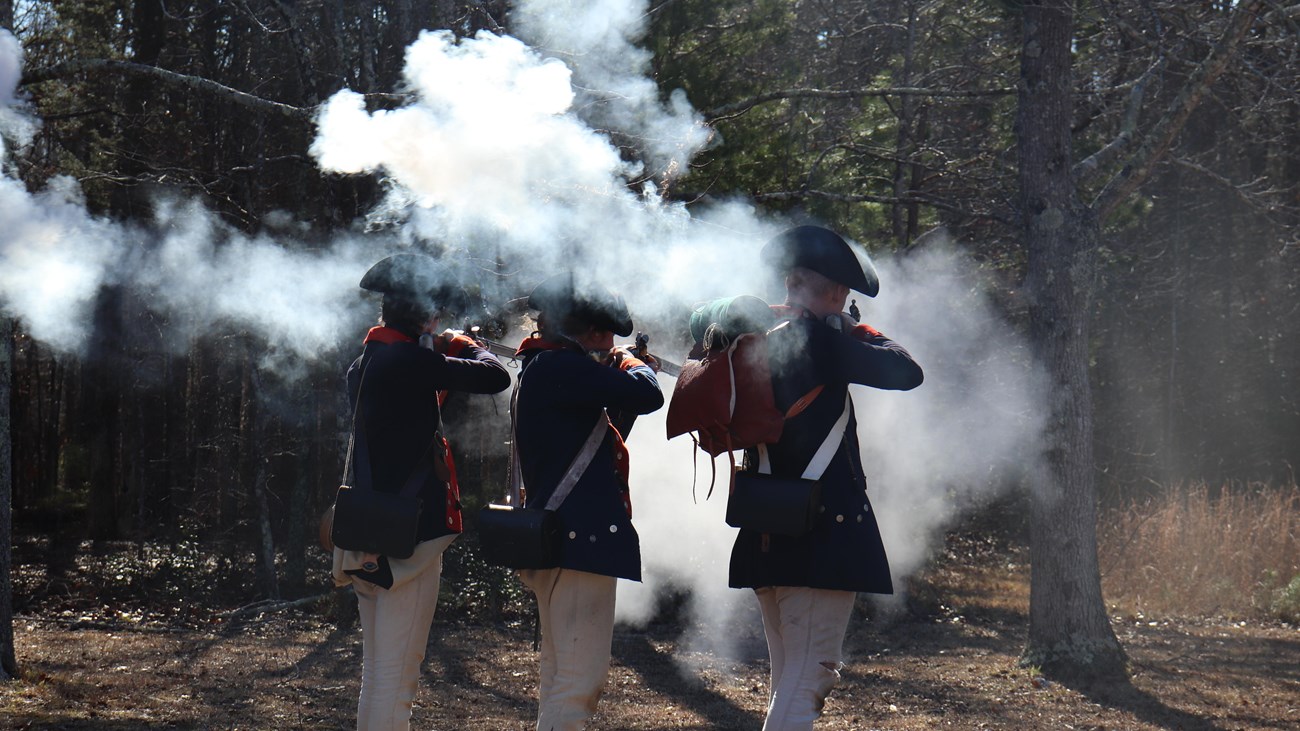 Three reenactors fire muskets in a cloud of smoke