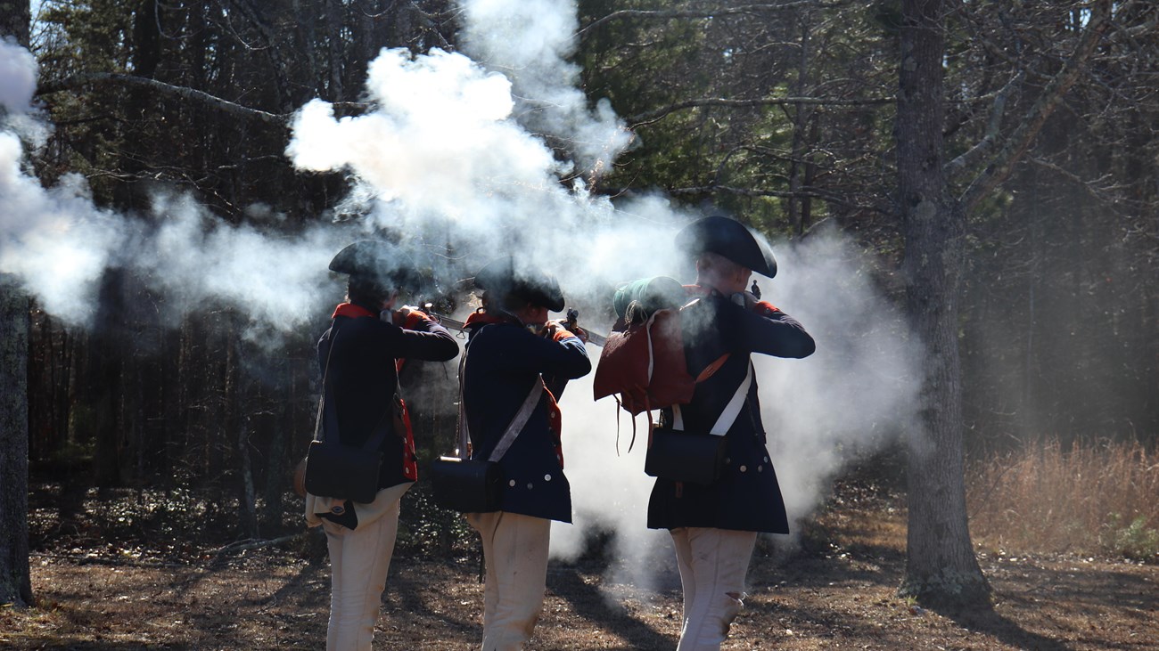 Three people in blue coats stand holding muskets surrounded by smoke 