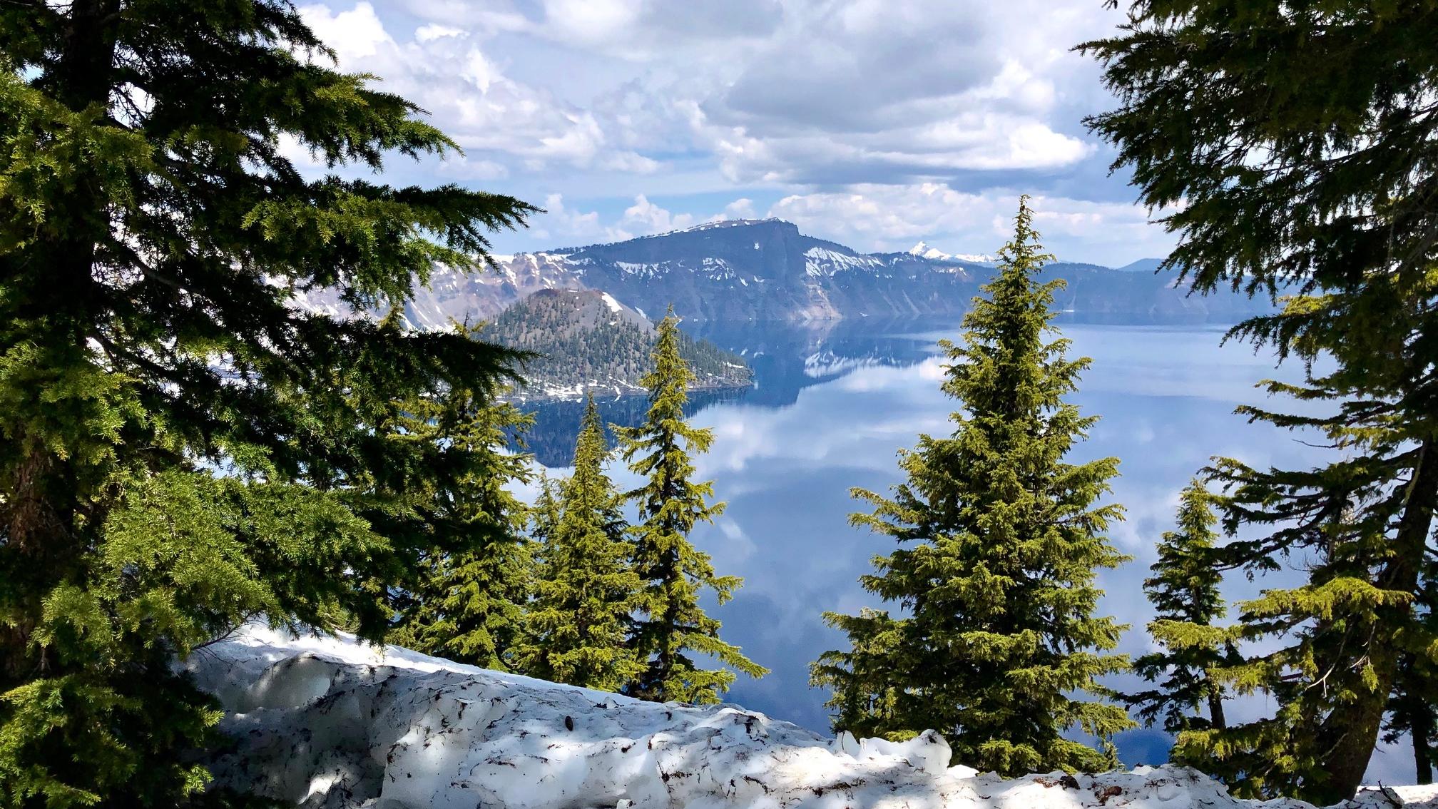 A mostly cloudy day, reflections of clouds in the lake, snow lingering around the caldera rim. 