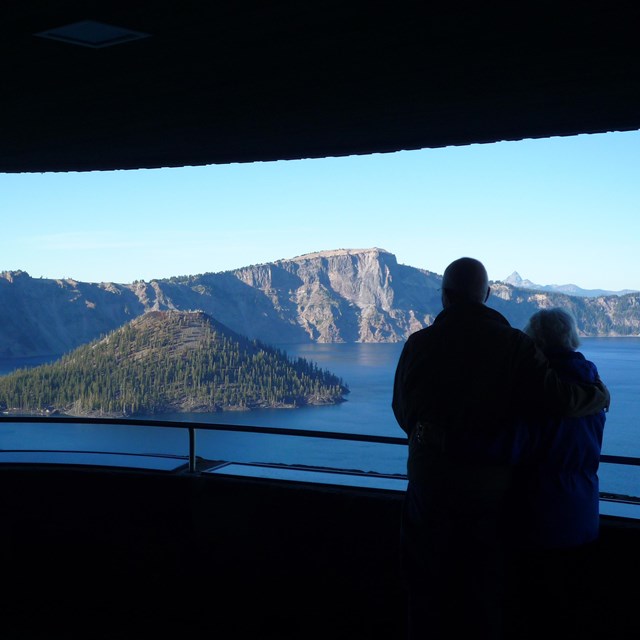 The silhouette of two visitors in a round stone shelter, looking across the lake and Wizard Island