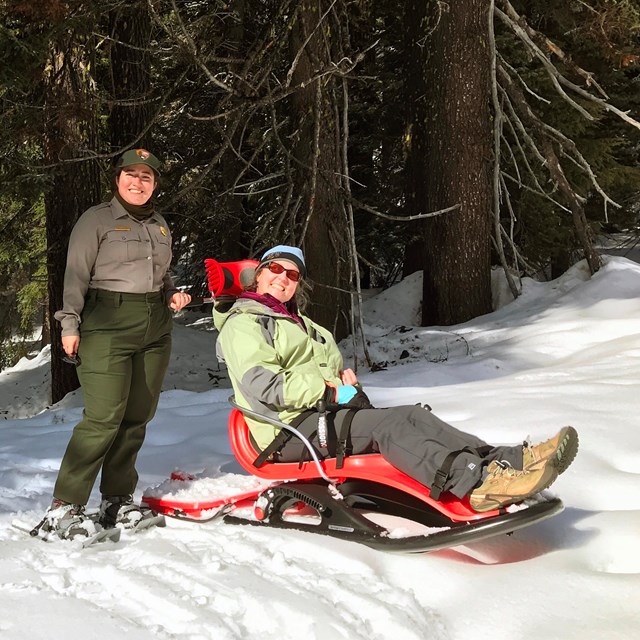 female park ranger on snowshoes and a female visitor sits in an adaptive sled on a snow field 