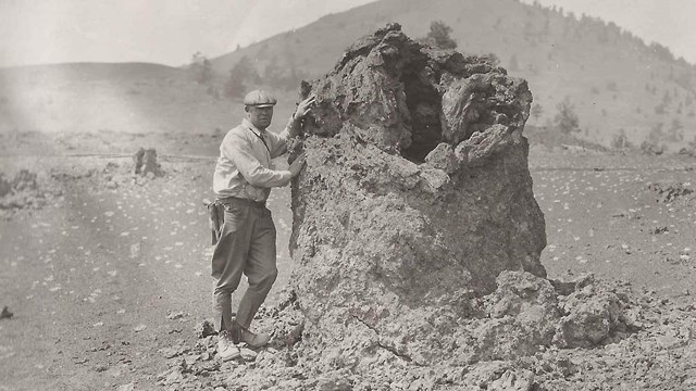 black and white photo of a man standing next to a lava formation slightly taller than him