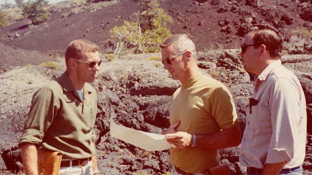photo of three men talking and looking at a piece of paper in front of dark lava rocks