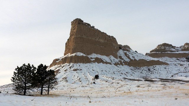 a tall, steep-sided rock formation standing alone above a flat, snowy landscape