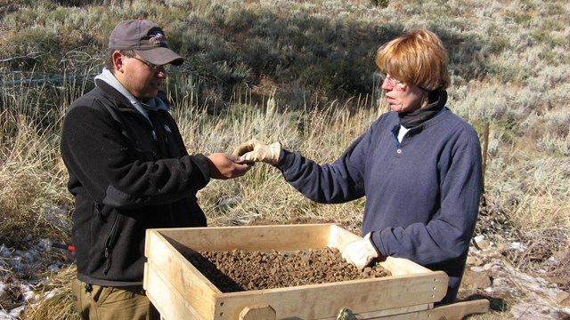 two people standing at a sifting crate and examining a small unknown object