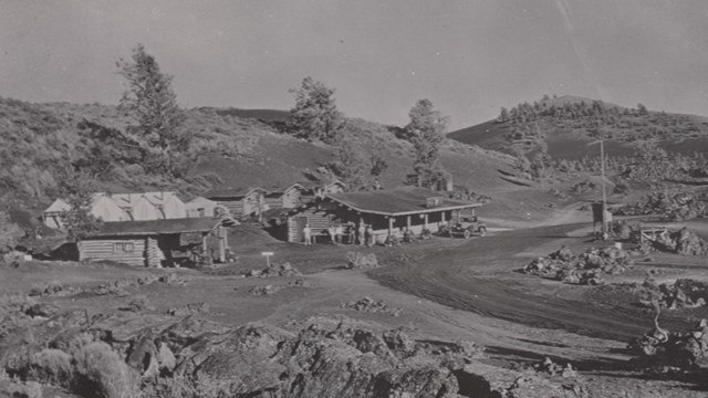black and white photo of two log buildings and several white tents amidst black rocks and cinders