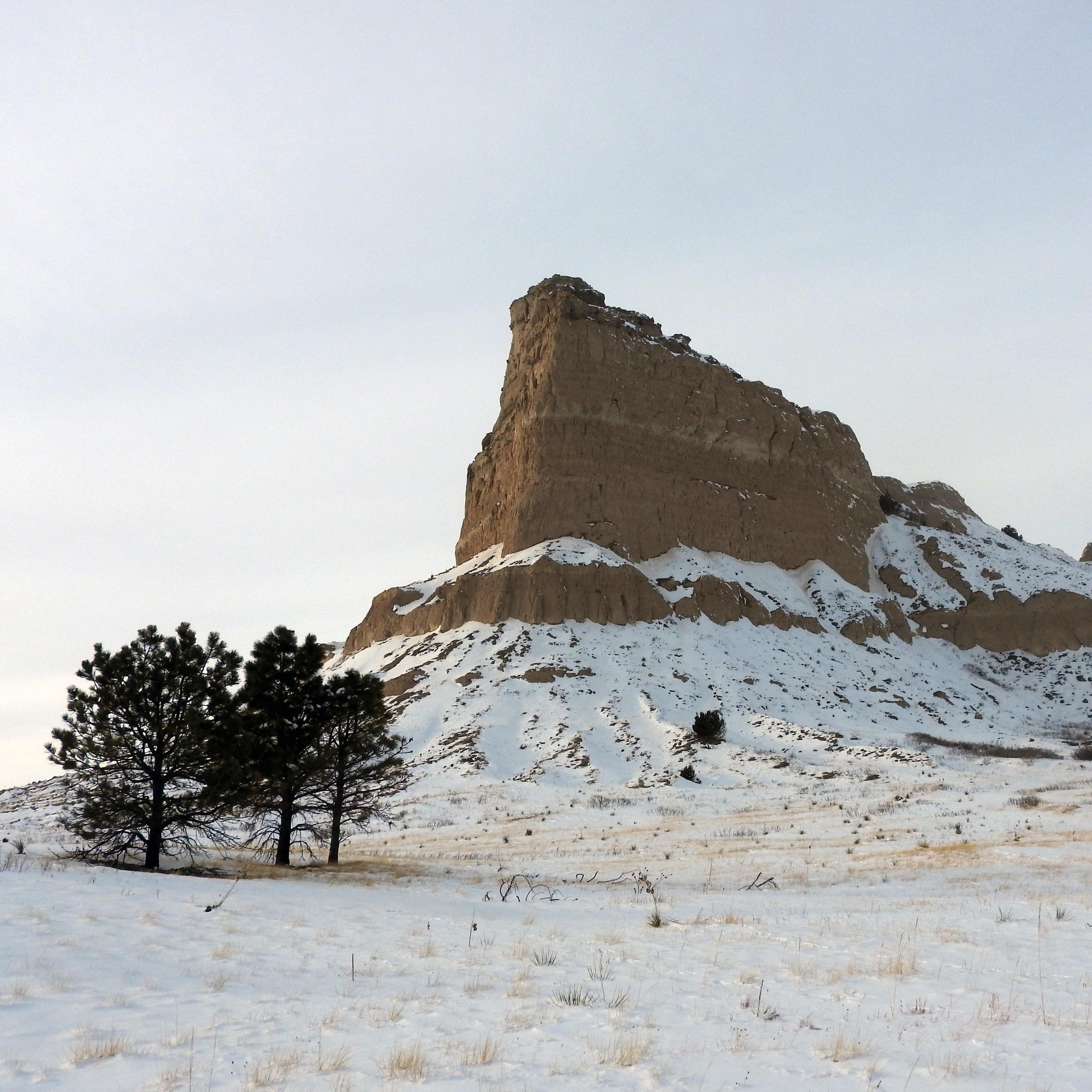 a tall, steep-sided rock formation standing alone above a flat, snowy landscape