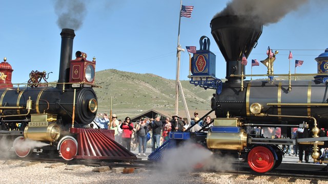Two 19th-century steam engines facing each other on a railroad near a crowd