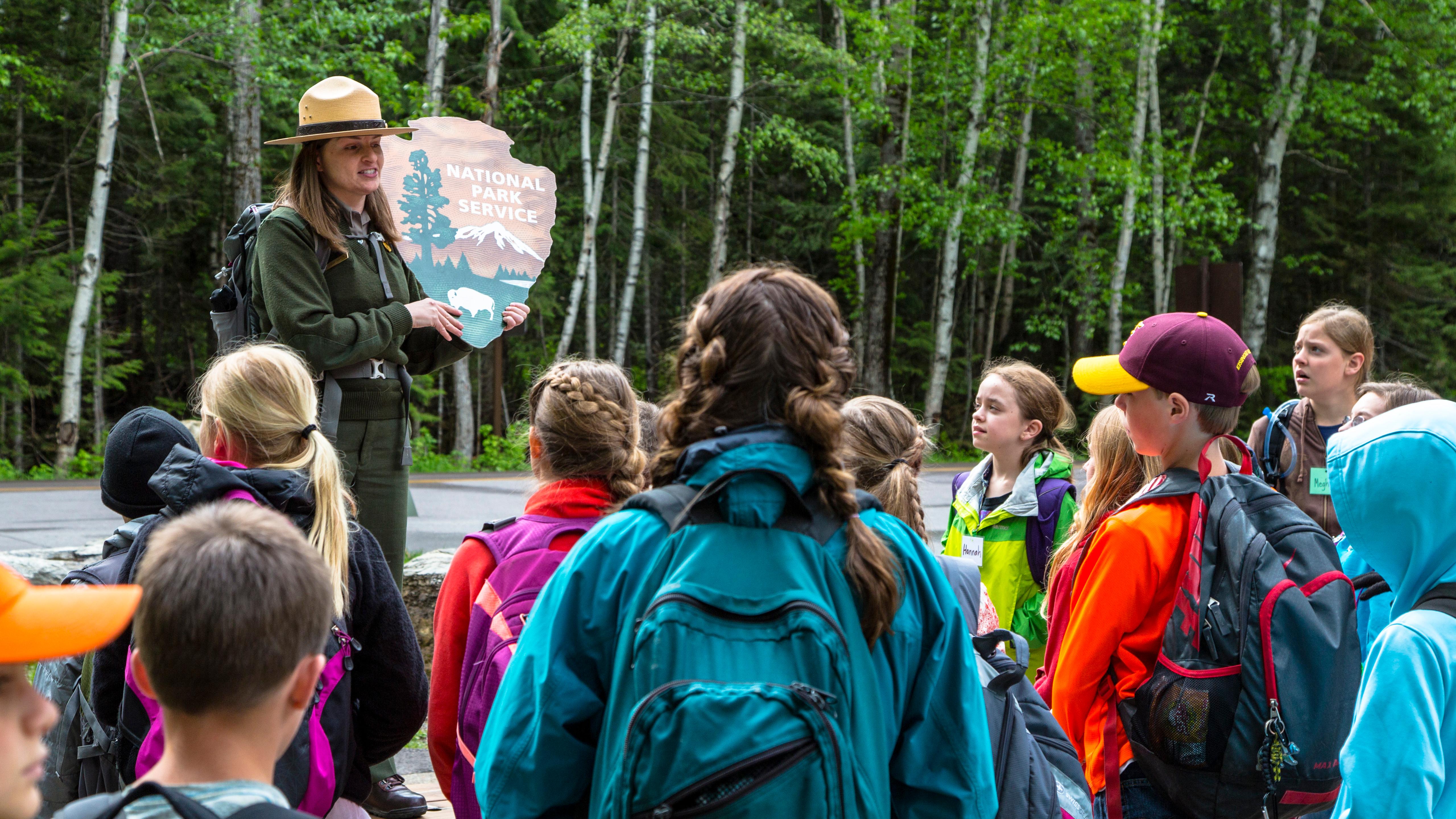 Ranger holding an NPS arrowhead logo while talking to a school group outside