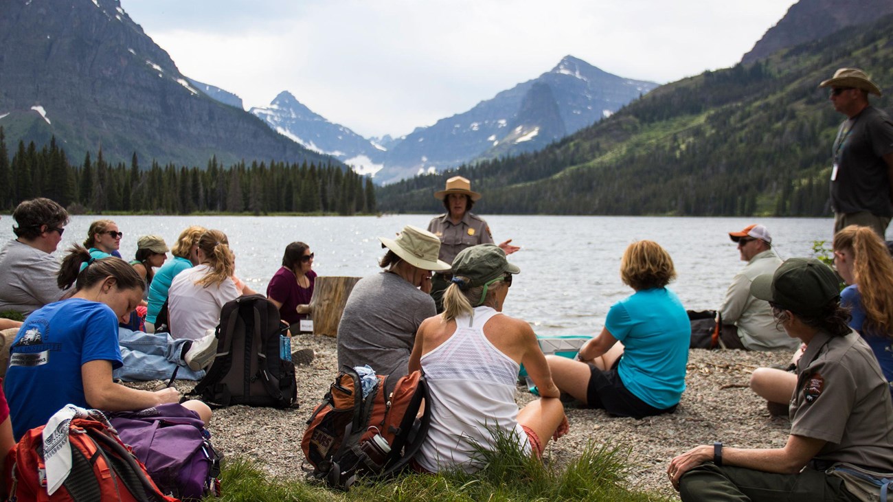 Teachers sit on the lakeshore of a mountain lake and listen to a park ranger.