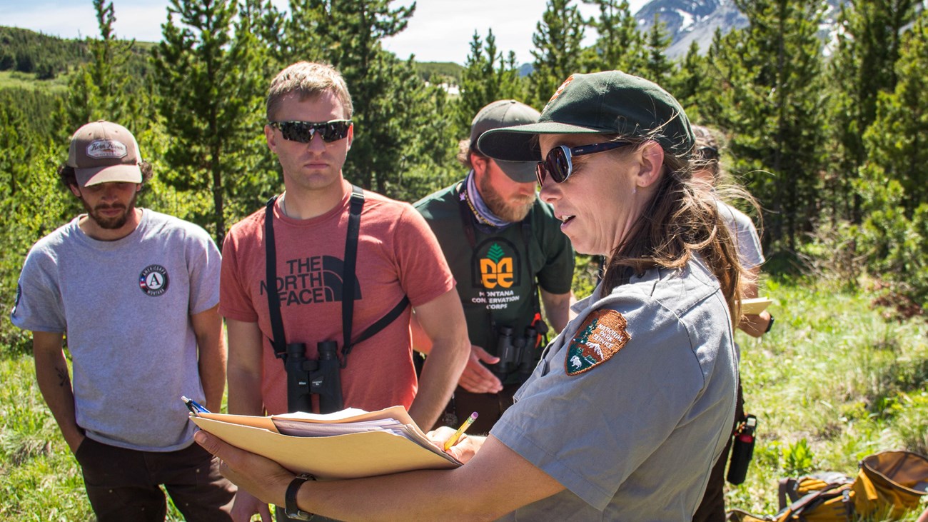 Out in a meadow, a woman in NPS uniform holds a clipboard and speaks to others standing nearby.
