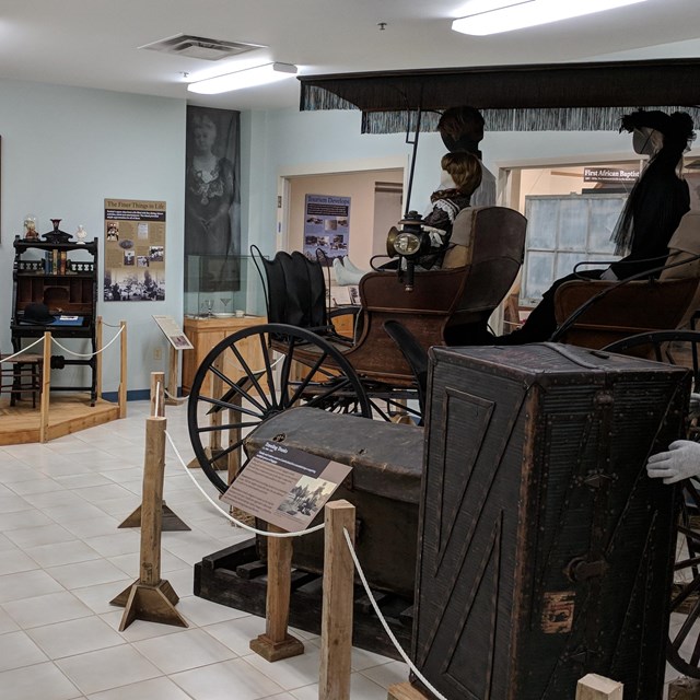 Horse drawn carriage, traveling trunks, and furniture, displayed with mannequins in period dress