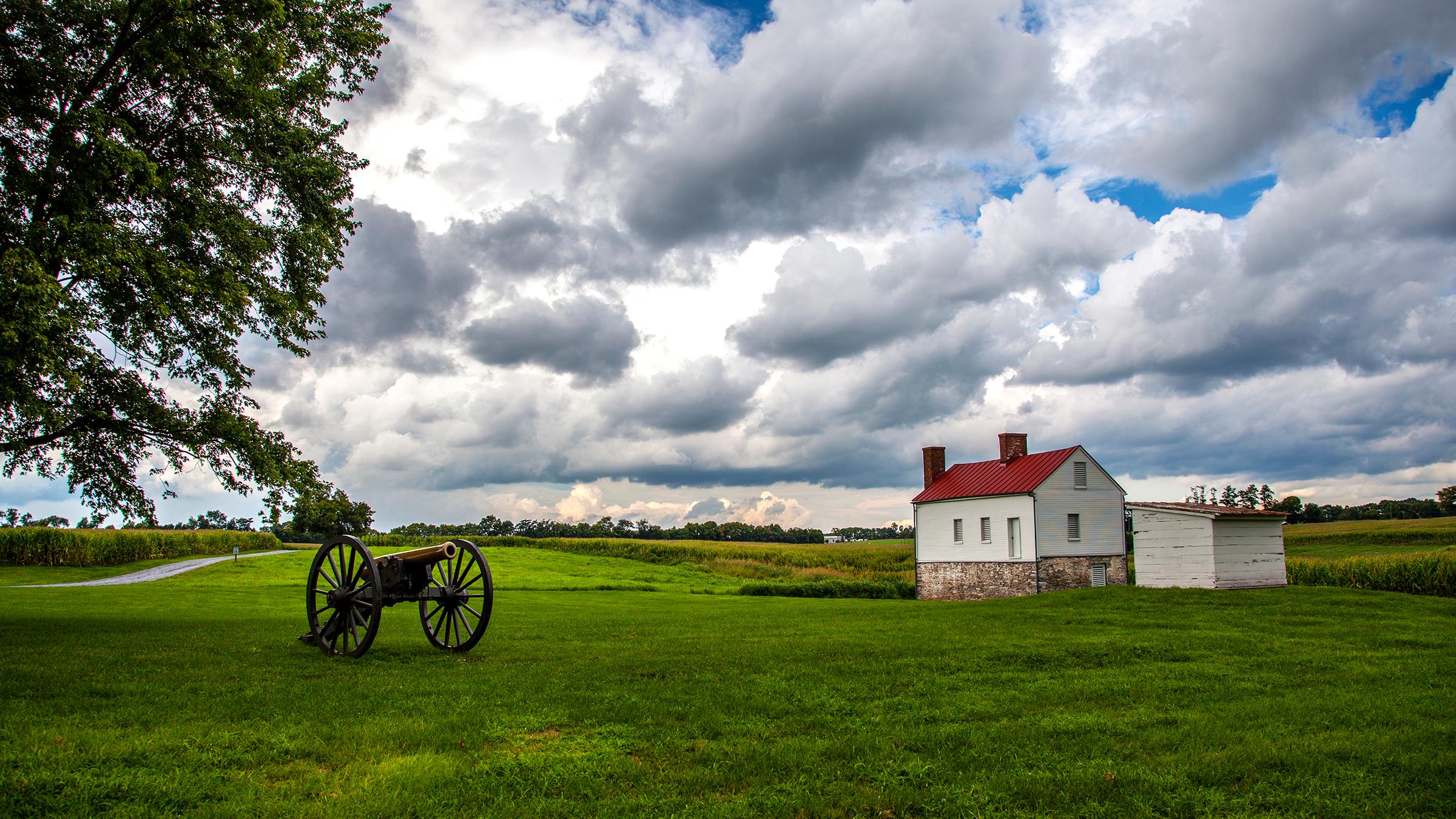 Puffy clouds hang over a red-roofed farm building and a cannon, surrounded by green grass. 