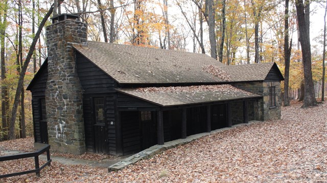 A one and a half story cabin in autumn woods has wooden siding, a stone chimney, and covered porch.