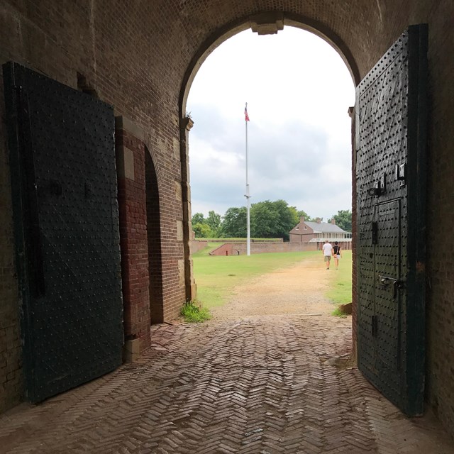 A view through an arched opening with a heavy door reveals a flag and parade ground.