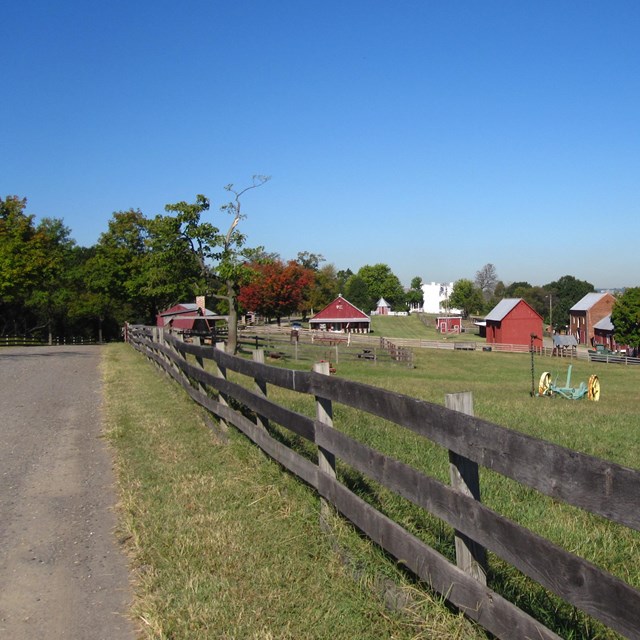 A wooden fence runs along a road that leads towards a cluster of farm buildings.