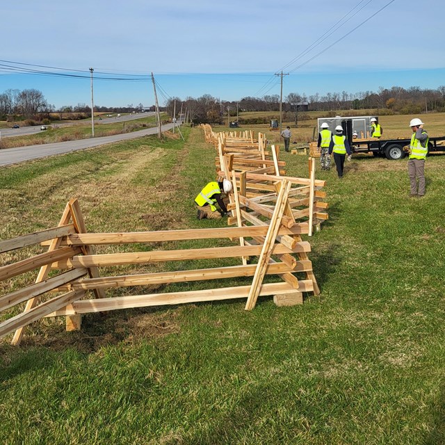 People in bright vests and hard hats work on a wooden splitrail fence near a road