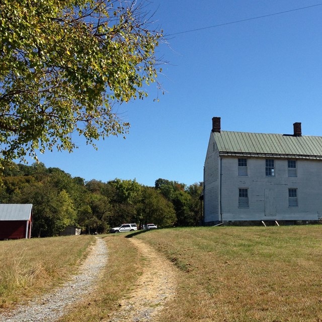 A dirt driveway curves towards a two-story farm house on a grassy expanse, under clear blue sky.