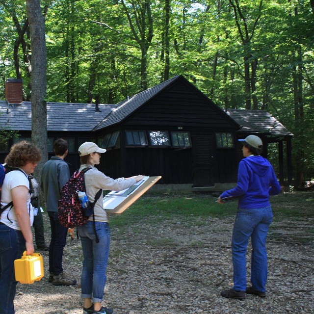 A group of people with documentation gear and site plans stand in a wooded area beside a cabin