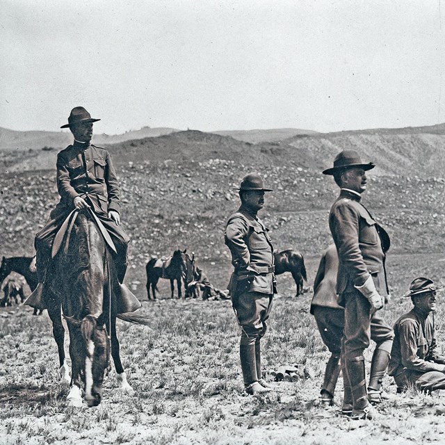 Soldiers in uniform and flathats gather in a field at Yellowstone, one on a horse.