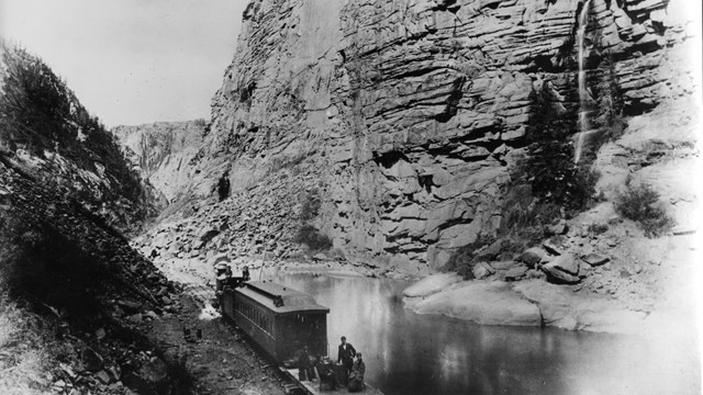 Historic image of a train on railroad tracks in a canyon. A low river is beside the tracks.