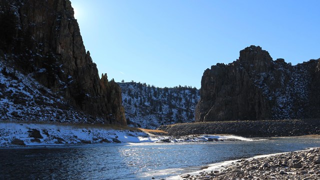 A river with snow and ice flows between two dark canyon walls. Blue sky is above the river.