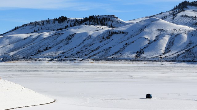 A small ice fishing tent is on a large frozen body of water. Snowy mesas in the background.