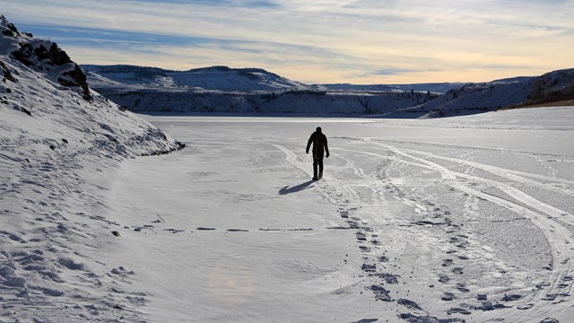 Silhouetted person snowshoes on a frozen snow-covered reservoir. Clouds and sunlight above.