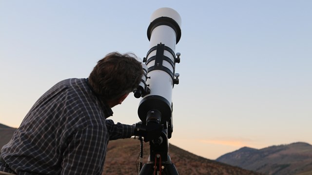 A person stands behind a large white telescope pointed towards a sky at dusk.