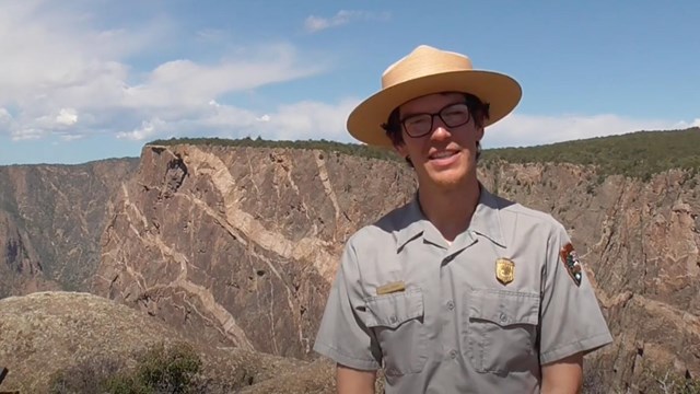 A person wearing a grey shirt and straw ranger hat stands in front of a canyon