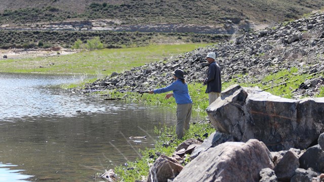 Two people casting fishing lines from the rocky shoreline of a lake