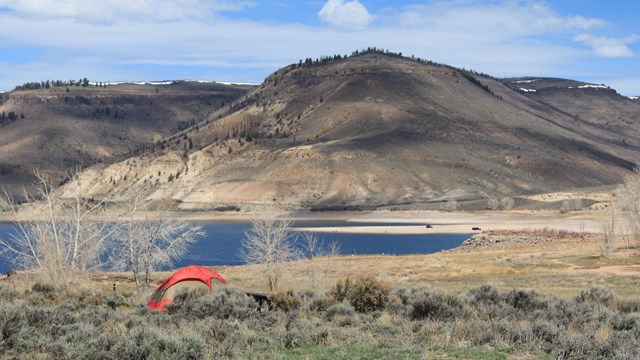 A small red tent pitched in a sagebrush area. A blue reservoir and vehicles are visible far off.