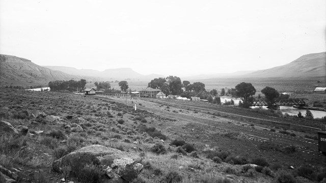 Historic black and white image of a railroad depot, buildings, river, and mesas.