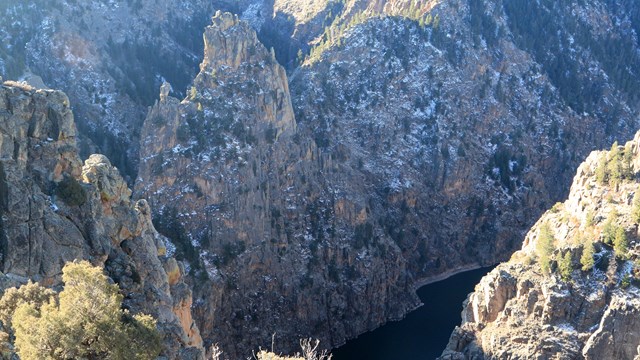 Tall canyon walls have snow in the shaded areas. A dark body of water is at the bottom.