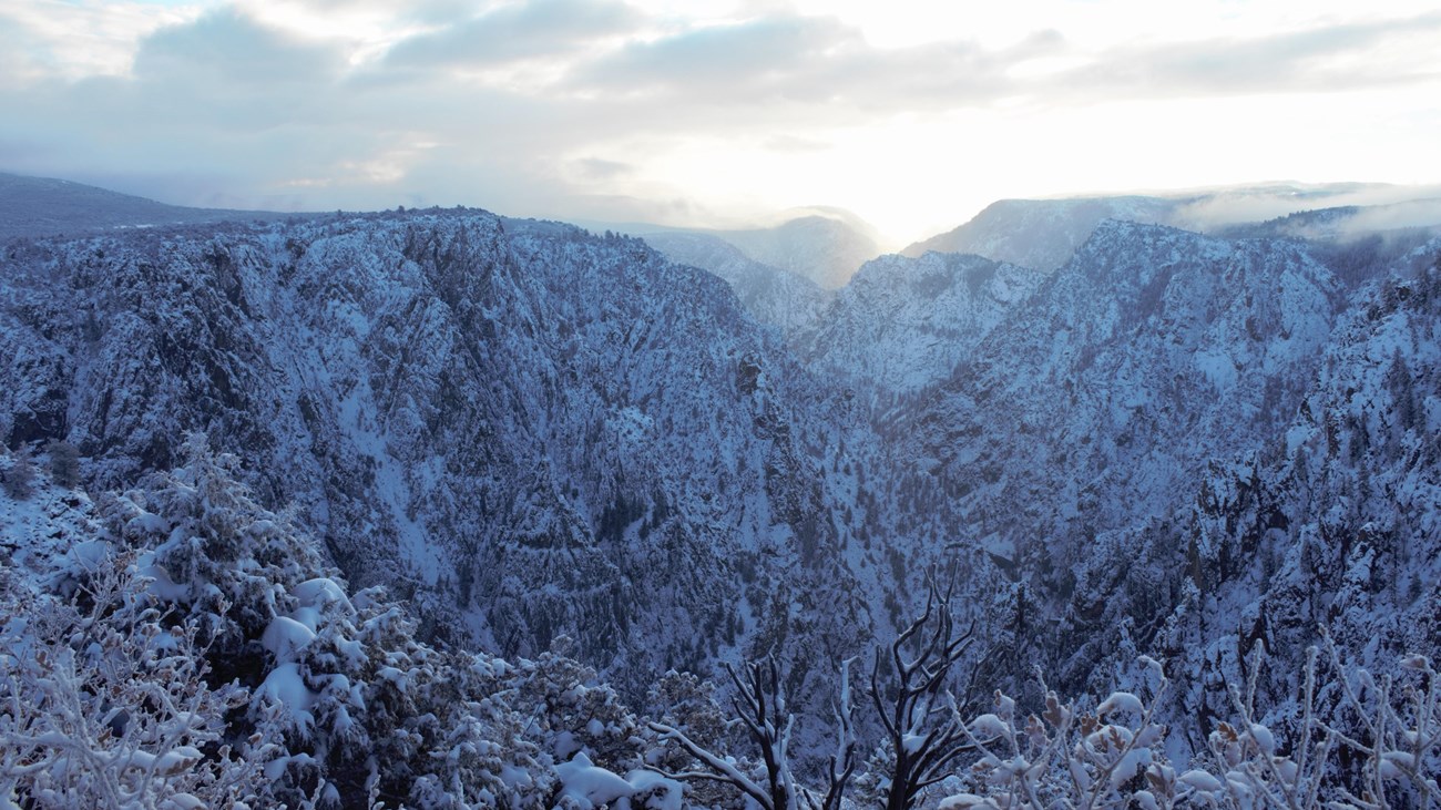 Sun illuminates snow along dark canyon walls. Blue sky and clouds are above.