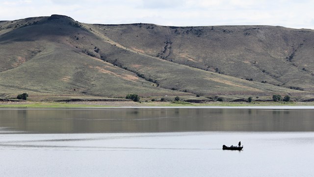 A person stands on the edge of a boat on a reservoir. Brownish-green mesas are in the background.
