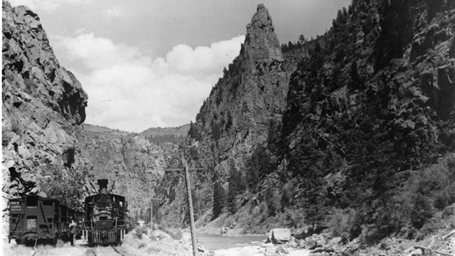 Historic image of a railroad line with a train between two steep canyon walls and a river.