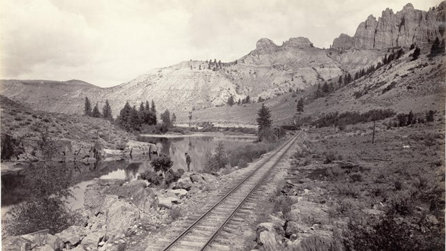 Historic image of a railroad, a man standing by a river, and mesas in the background.