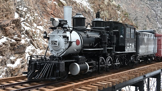 A black locomotive and other train cars sit on a metal and wooden truss bridge in a canyon