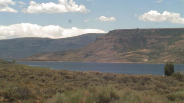 Screenshot of a webcam image showing brownish-grey mesas and large blue reservoir