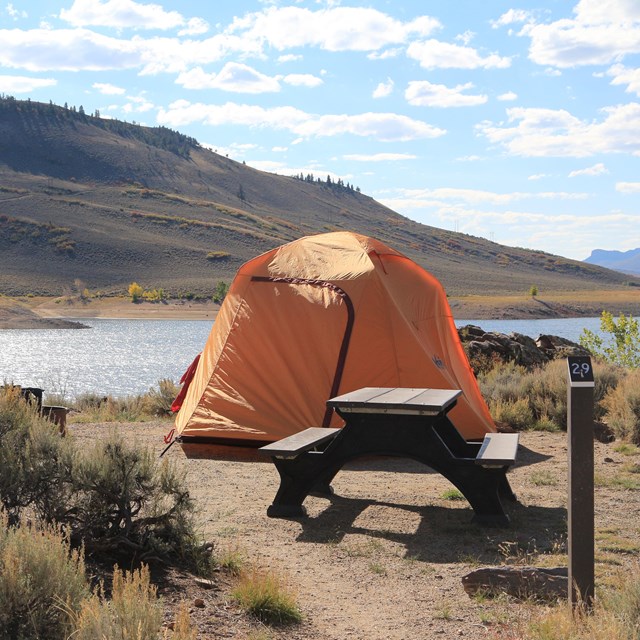 An orange tent is pitched behind a picnic table and fire grate. Sagebrush and water are behind it.