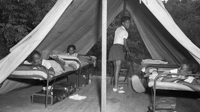 Four Black girls in a tent on a wooden platform; three lie in beds while the fourth uses a broom.