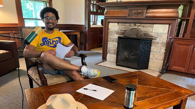A Black man with short afro and glasses sits behind a microphone by a fireplace.