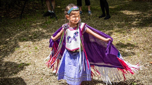 Young girl in a colorful dress stands smiling, her outstretched arms holding her fringed cape open.