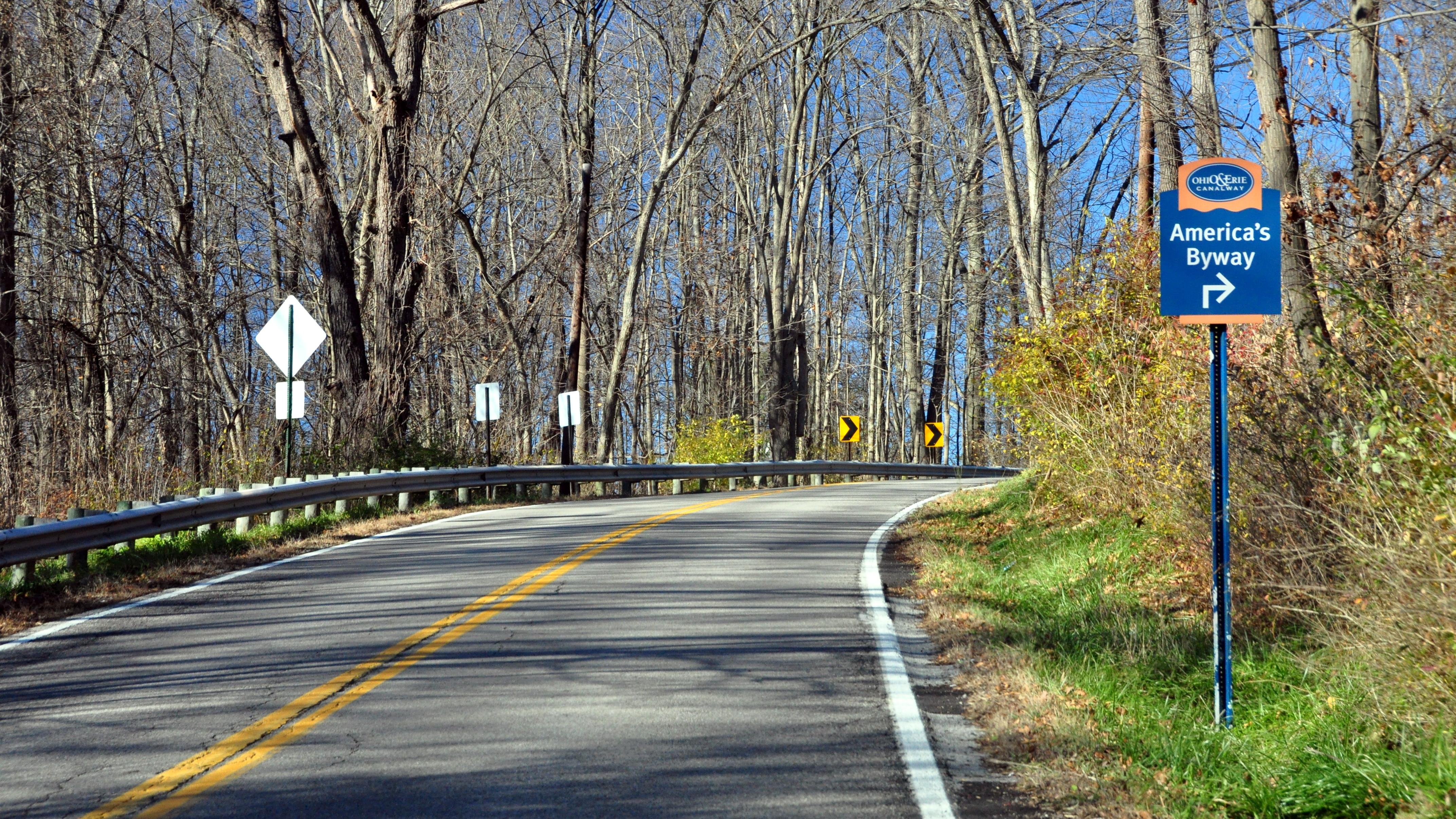A road curves through a sunny forested landscape; a blue sign to the right reads "America's Byway."