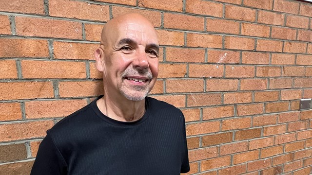 A smiling, bald man with a grey beard stands against a brick wall.