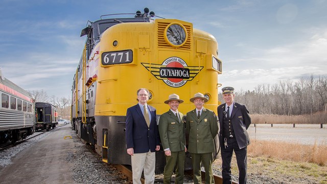Uniformed rangers and two others stand in front of a yellow Cuyahoga Valley Scenic Railroad train.